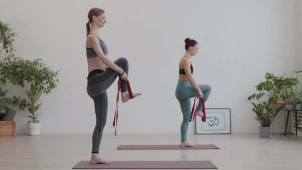 Широкий снимок двух молодых йогов в спортивной одежде, стоящих на одной ноге на ковриках для йоги в студии йоги и растягивающих другую ногу с помощью йогического пояса - Кадры, видео