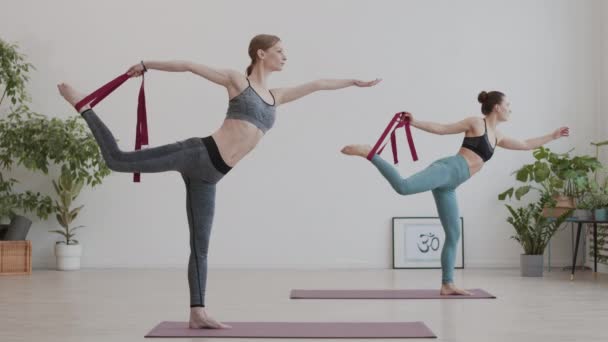 Amplio tiro de dos jóvenes deportistas con ropa deportiva de pie sobre una pierna en esteras de yoga en el gimnasio y estirando la otra pierna usando cinturón yóguico - Metraje, vídeo