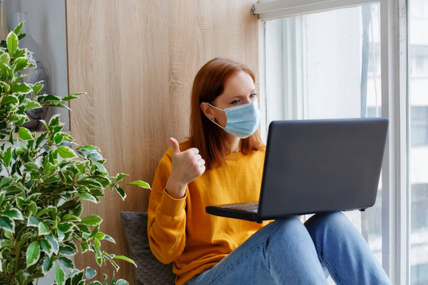 A female employee or student sits at the window with a mask on her face and a laptop. Cozy and convenient work remotely. - Фото, изображение