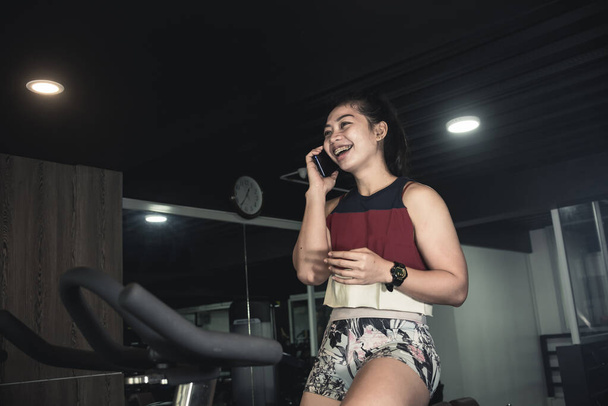 Una mujer asiática deportiva habla con su amigo por teléfono mientras está en la bicicleta estacionaria haciendo cardio. - Foto, Imagen