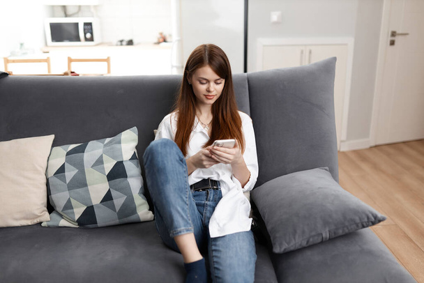 Μια γυναίκα στο σπίτι με ένα τηλέφωνο στα χέρια της αναπαύεται σε έναν άνετο καναπέ. - Φωτογραφία, εικόνα