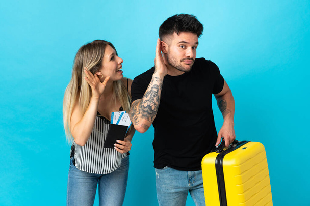 Young caucasian couple going to travel isolated on blue background listening to something by putting hand on the ear - Photo, image