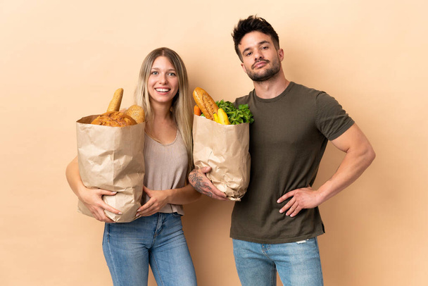 Casal segurando supermercado sacos de compras sobre fundo isolado posando com braços no quadril e sorrindo - Foto, Imagem