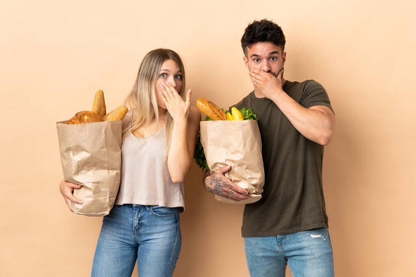 Casal segurando supermercado sacos de compras sobre fundo isolado cobrindo boca com as mãos para dizer algo inapropriado - Foto, Imagem