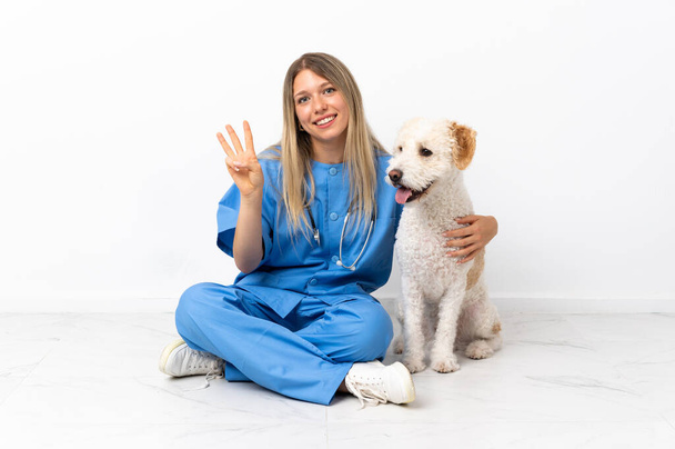 Young veterinarian woman with dog sitting on the floor smiling and showing victory sign - Zdjęcie, obraz