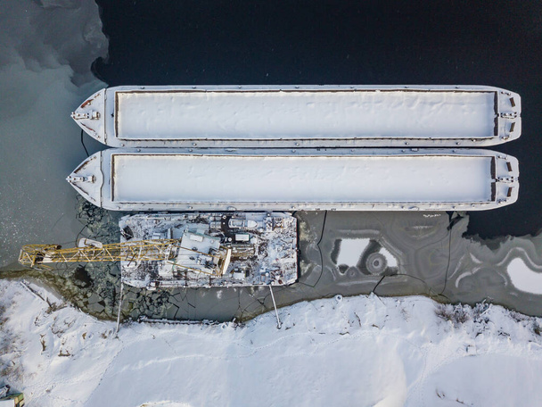 Ένας γερανός κοντά σε ένα φορτηγό πλοίο σε ένα παγωμένο κόλπο. Αεροφωτογραφία τηλεκατευθυνόμενου. Χειμερινό χιονισμένο πρωινό. - Φωτογραφία, εικόνα