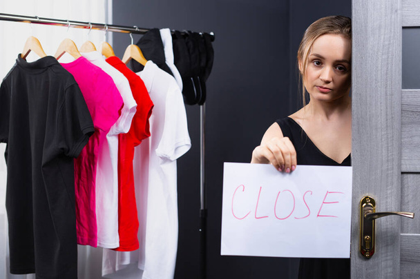 A kind and beautiful blonde, sad, woman working as a consultant in a clothing store indicates that the store is closed on a signboard. Photo indoor shop. High quality photo - Photo, image