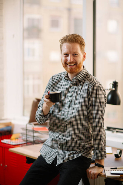 Красивый бизнесмен с рыжими волосами пьет кофе в офисе - Фото, изображение