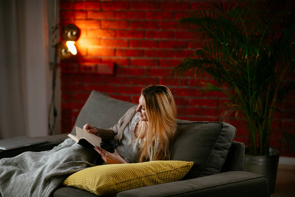 Evdeki kanepede kitap okuyan güzel bir kız. Rahat bir dairede kitap okuyan bir kadın.. - Fotoğraf, Görsel
