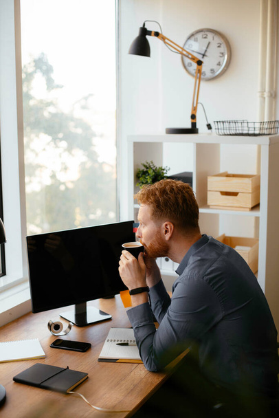 όμορφος επιχειρηματίας με κόκκινα μαλλιά πίνοντας καφέ στο γραφείο - Φωτογραφία, εικόνα