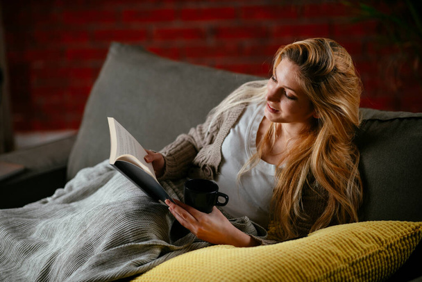 Young beautiful girl reading a book on the couch at home. Woman reading a book at cozy apartment. - Foto, imagen