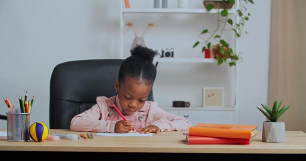 Kleine lustige Vorschulmädchen kreative talentierte afroamerikanische Kind sitzt allein am Tisch zu Hause zeichnet Bild auf Papier mit Buntstiften in Hobby am Wochenende Studie beschäftigt Hausaufgaben  - Filmmaterial, Video