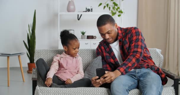 El padre afroamericano joven se sienta con su amada hija mostrando su video en el teléfono, la familia viendo películas de terror en dispositivos móviles, los golpes de niños que se esconden del miedo, el padre y la niña pasan tiempo en casa - Metraje, vídeo
