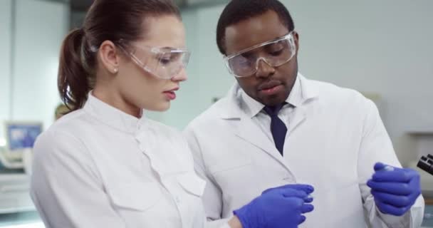 Detailní záběr smíšených ras mladých lékařů ženského a mužského pohlaví specializovaných na bílé pláště, kteří stojí v klinické laboratoři v brýlích a dělají krevní testy, diskutují o vzorcích, mikrobiologii. Koncept analýzy - Záběry, video