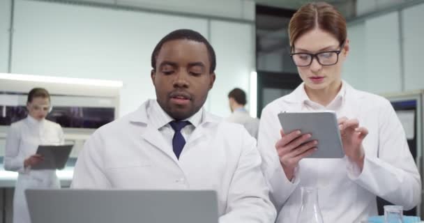 アフリカ系アメリカ人の若い男性医師が、ノートパソコンやタブレット上でのコロナウイルスワクチンの開発について診療所で働く女性科学者と話す様子 - 映像、動画