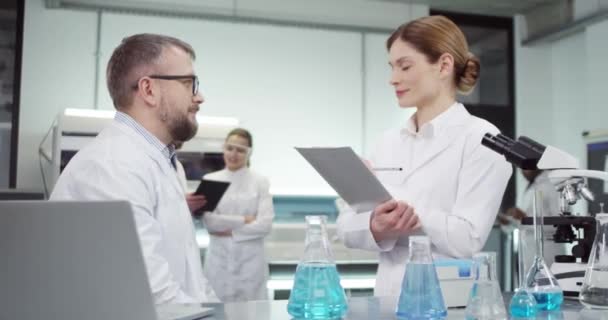 Portrait of pretty Caucasian female medical assistant speaking with middle-aged experienced male physician who is browsing on laptop working in clinic with different tubes with liquid on table - Footage, Video
