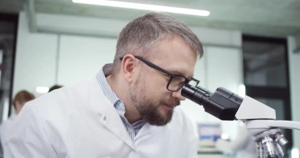 Κοντινό πορτρέτο του καυκάσιου γενειοφόρου μεσήλικα άνδρα γιατρού σε γυαλιά που εργάζεται στο εργαστήριο σε εξοπλισμό μικροσκοπίου που διεξάγει πειράματα ανάπτυξης εμβολίου covid-19 και δακτυλογράφησης σε υπολογιστή - Πλάνα, βίντεο