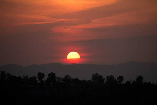 木々との典型的な日没アフリカの風景。大きなオレンジの丸みを帯びた夕日に対する木のシルエット。オープンフィールド劇的な日の出に暗い木。雲と夜明けの空、夕暮れの背景. - 写真・画像