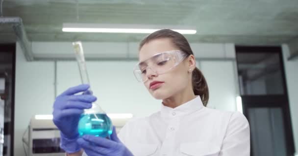Primo piano di gioiosa bella caucasica giovane scienziata di laboratorio donna in occhiali che lavorano sul vaccino covid-19 in laboratorio tenendo becher con sostanze chimiche liquide blu guardando concetto farmaceutico fotocamera - Filmati, video