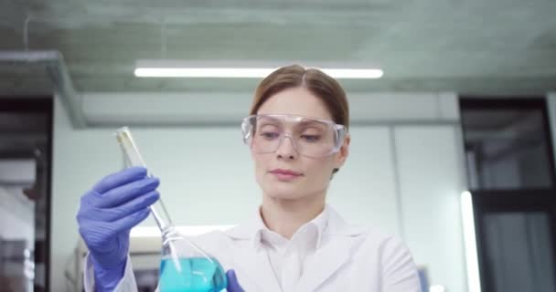 Großaufnahme der schönen kaukasischen erfahrenen Laborantin mit Brille, die im Labor in der Klinik arbeitet und einen Becher mit blauem Flüssigchemikalienimpfstoff in die Kamera hält und lächelt - Filmmaterial, Video
