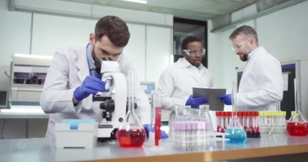 Kaukasische beschäftigte männliche Arzt Spezialist im Labor unter dem Mikroskop untersucht DNA-Mutationen, die Entwicklung von Virus-Impfstoff. Ein afroamerikanischer Arzt spricht mit einem Kollegen im Hintergrund. Porträt - Filmmaterial, Video