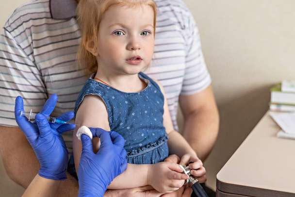 Маленькая девочка с отцом в кабинете врача в клинике прививается от коронавируса. Концепция вакцинации, иммунизации, профилактики против вируса Ковид-19 - Фото, изображение
