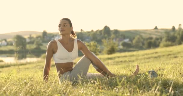 Caucasian beautiful young woman sitting on green grass and turning back to stretch side, hip and back. Pretty sporty female yogi stretching early in the morning. Physical training at nature concept. - Footage, Video
