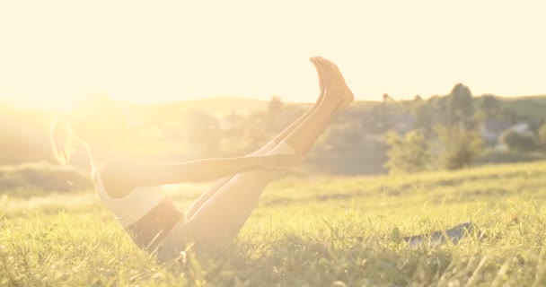 Csinos fiatal sportos nő jógi ül a zöld füvön és egyensúlyozik felemelt kézzel és lábakkal. Napfényben. Női kezdő nyári nap testmozgással és jógával. Bájos természet. Nyújtott ászana. - Felvétel, videó