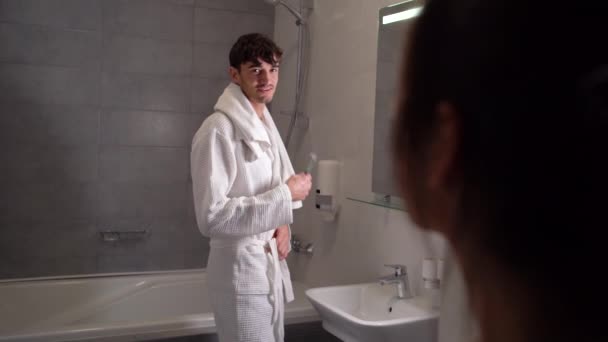 Een knappe jonge brunette blanke man in een witte jas poetst tanden in de badkamer terwijl zijn vriendin met hem praat vanaf de deur. - Video
