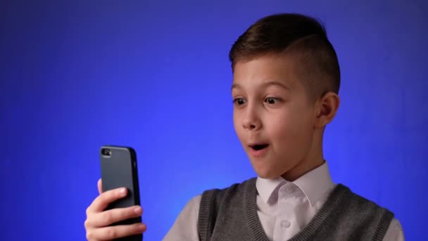 Bellissimo ragazzo della scuola guarda è sorpreso a smart phone su sfondo blu. Bambino moderno è la gioia guardando gadget. - Filmati, video
