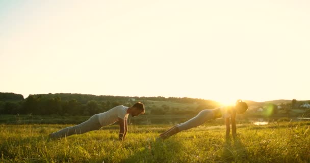 Caucasiano masculino e feminino yogis de pé na prancha na grama verde para baixo com belo sol e natureza no fundo. Homem e mulher fazendo ioga de manhã cedo. - Filmagem, Vídeo