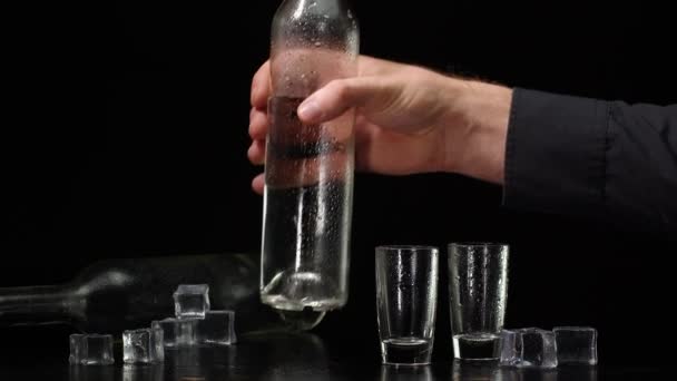 Cantinero vertiendo vodka de botella en dos vasos de chupito con cubitos de hielo sobre fondo negro - Imágenes, Vídeo