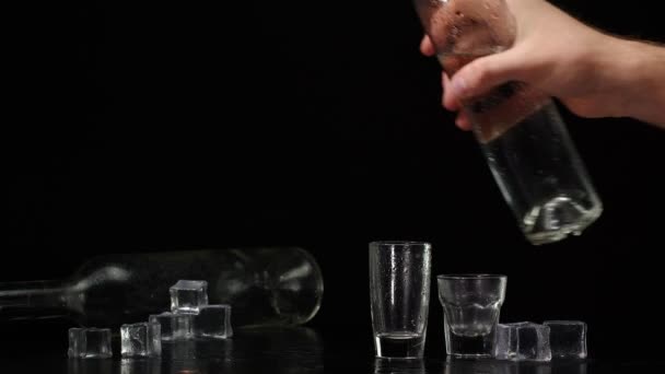 Bartender pouring up two shots of vodka with ice cubes from bottle into glasses on black background - Footage, Video