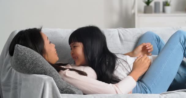 Щасливий безтурботний азіатська корейська мати і маленька дитина дочка лежить на дивані диван сміється весело посміхаючись разом, дитина цілує маму на носі, насолоджуючись зв'язуванням обіймів вдома у вихідні
  - Кадри, відео