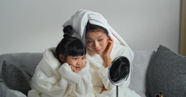 Pareja de bellezas joven madre y pequeña hija asiática familia usar batas de baño y toallas en sus cabezas mirada en reflejo en espejo madre alisa sus hijos peinado preen sentado en casa - Metraje, vídeo