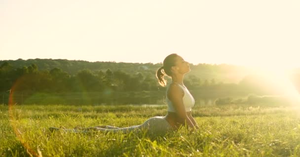 Biała młoda kobieta jogin leży na zielonej trawie, a następnie rośnie w pozycji kobry medytować i oddychać. Piękna wiejska przyroda. Sportowa kobieta w yoga asana zaczyna letni dzień wcześnie rano. Na zewnątrz. - Materiał filmowy, wideo