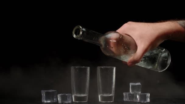 Cantinero vertiendo vodka de botella en dos vasos de chupito con cubitos de hielo sobre fondo negro - Imágenes, Vídeo