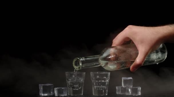 Barman nalewa dwa kieliszki wódki z kostkami lodu z butelki do szklanek na czarnym tle - Materiał filmowy, wideo