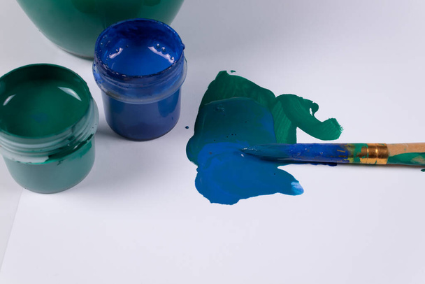 zwei offene Gläser Gouache nebeneinander und auf einer weißen Fläche neben ein paar Strichen Grün und Blau, und eine schmutzige Bürste liegt darauf, neben einem Glas Wasser - Foto, Bild