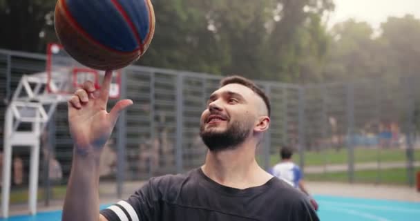 Close up portrait of joyful Caucasian basketball player standing on street and spinning ball on finger outdoors. Cheerful athlete resting on playground while friends playing streetball. Sport concept - Footage, Video