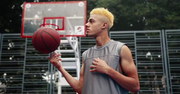 Портрет серьезного молодого красивого спортсмена с светлыми волосами, стоящего на баскетбольной площадке под открытым небом и бросающего мяч. Сконцентрированный игрок в стритбол смотрит в сторону города. Концепция уличного спорта - Кадры, видео