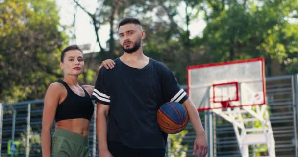 Portré vidám fiatal férfi és női sportolók álló szabadtéri kosárlabda játszótér. Kaukázusi gyönyörű boldog lány és jóképű fiú golyóval pózol a kamerába. Atlétikai koncepció - Felvétel, videó