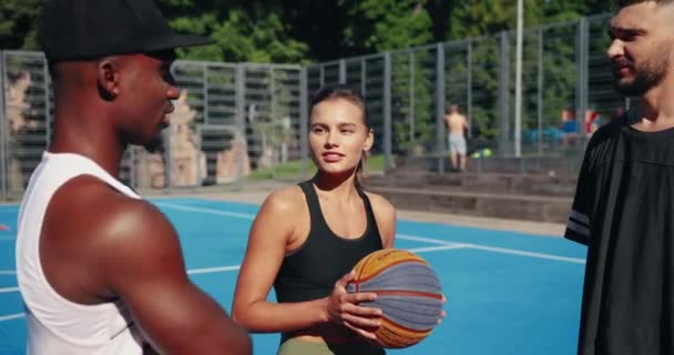 Close up portrait of mixed-races male and female basketball players standing on outdoor court and talking. Beautiful Caucasian girl and guy speaking with handsome African American man about game - Footage, Video