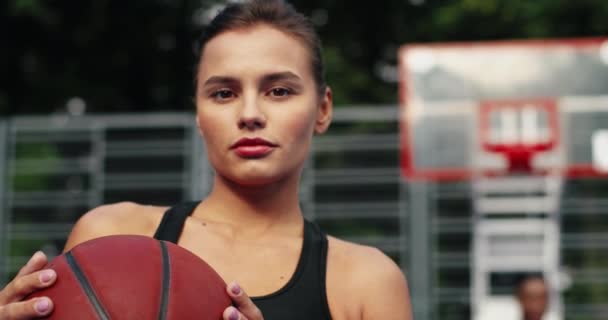 Közelkép komoly, fiatal, csinos, kaukázusi női atléta kosárlabdára néző kamerával, miközben kint áll a játszótéren. Gyönyörű, nyugodt arcú nő portréja. Atlétikai koncepció - Felvétel, videó