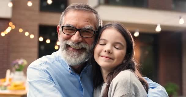 Portrait of Caucasian grandfather in glasses hugging small cute granddaughter and smiling in yard. Old gray-haired man embracing little pretty girl. Celebration outdoors. Family dinner and meeting. - Footage, Video