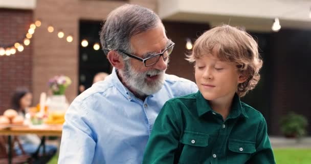 Portrait of Caucasian grandfather in glasses hugging small cute grandson and smiling in yard. Old gray-haired man embracing little boy. Celebration outdoors. Family dinner and meeting. Happiness. - Footage, Video