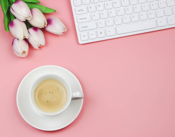 Bovenaanzicht of platte lay-out van koffiebeker met tulpenbloem boeket en computer toetsenbord op roze achtergrond met kopieerruimte voor tekst. Vrouwelijke werkplek. Ochtendroutine. - Foto, afbeelding
