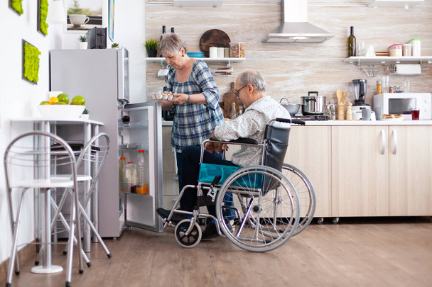 Старша жінка готує сніданок для чоловіка з інвалідністю, який приймає яйця з холодильника
 - Фото, зображення