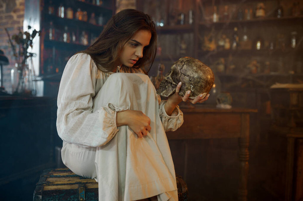 Straszna demoniczna kobieta trzyma ludzką czaszkę, demony wypędzają. Egzorcyzmy, tajemniczy paranormalny rytuał, ciemna religia, nocny horror, mikstury na półce w tle - Zdjęcie, obraz