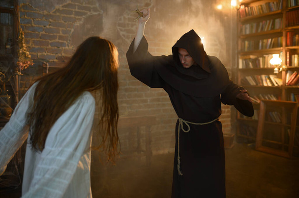 Male exorcist in black hood casting out satan from crazy woman. Exorcism, mystery paranormal ritual, dark religion, night horror, potions on shelf on background - Photo, Image